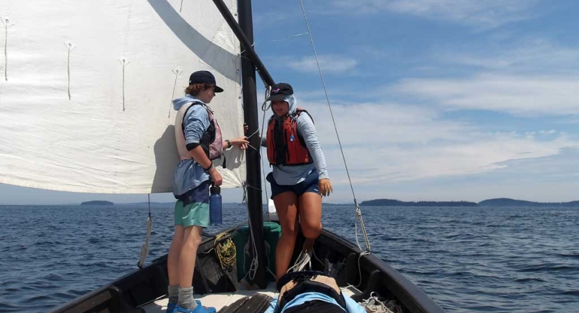 Two people stand at the front of a sailboat near a white sail. 